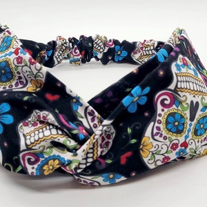 May include: A black headband with a white, pink, blue, and yellow sugar skull pattern. The headband is made of a soft, stretchy fabric and is designed to be worn as a hair accessory.