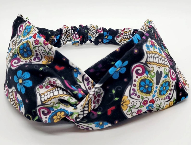 May include: A black headband with a white, pink, blue, and yellow sugar skull pattern. The headband is made of a soft, stretchy fabric and is designed to be worn as a hair accessory.