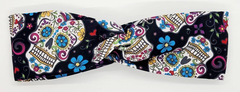 May include: A black headband with a white and colorful sugar skull pattern. The headband is twisted and has a knot in the center.