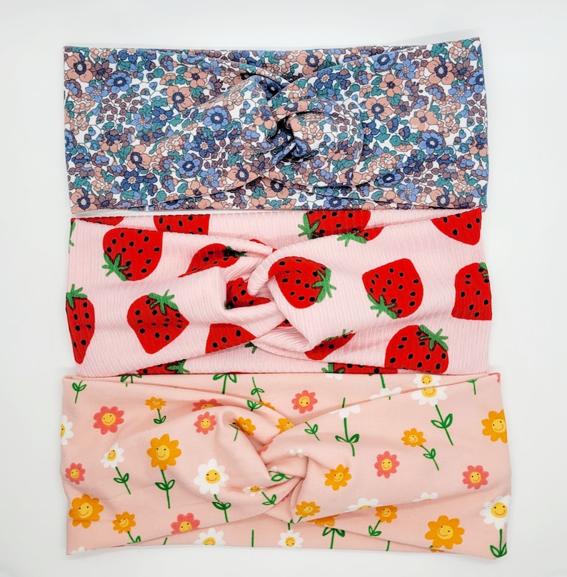 May include: Three pink fabric headbands with different patterns. The top headband has a floral pattern in shades of blue, pink, and white. The middle headband has a pink background with red strawberries. The bottom headband has a pink background with yellow smiley faces.