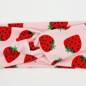 May include: Pink stretchy headband with a knotted design and a repeating pattern of red strawberries on a white background.