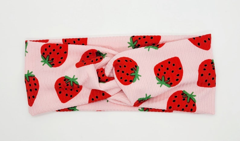 May include: Pink stretchy headband with a knotted design and a repeating pattern of red strawberries on a white background.