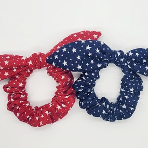 May include: Two scrunchies with star patterns. One is red with white stars, and the other is navy blue with white stars. Each scrunchie has a bow-like detail.