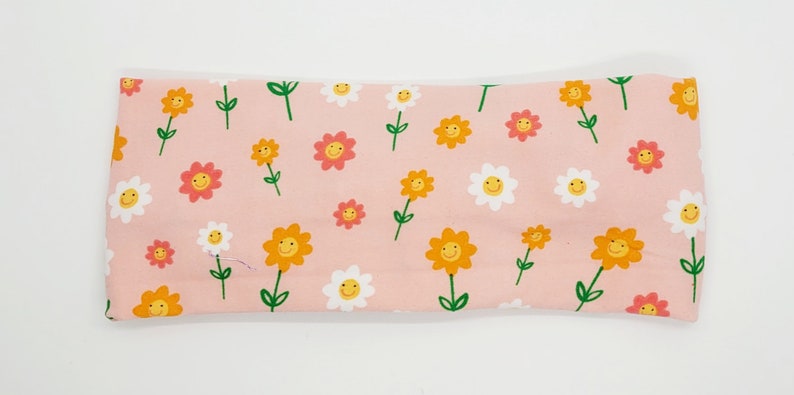 May include: A pink headband with a repeating pattern of yellow, orange, and white flowers with smiley faces.