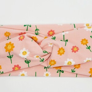 May include: A pink headband with a pattern of white and yellow flowers with smiley faces. The headband is twisted and has a knotted design.
