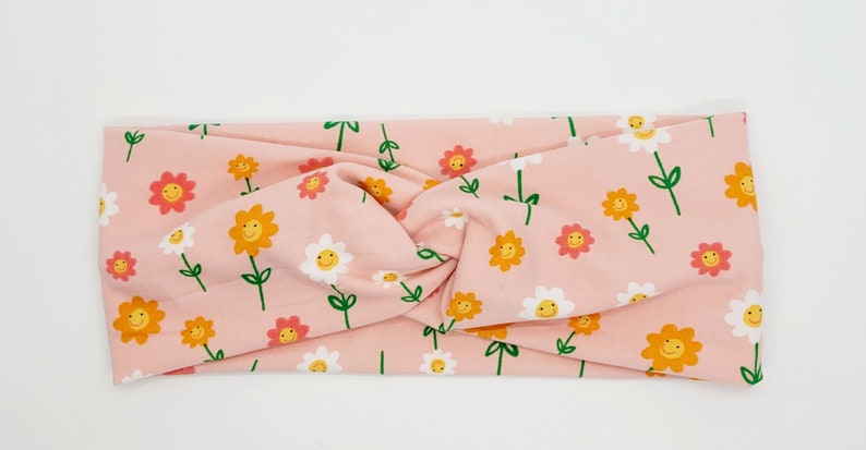 May include: A pink headband with a pattern of white and yellow flowers with smiley faces. The headband is twisted and has a knotted design.