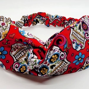 May include: Red headband with a white and colorful sugar skull pattern. The headband is made of a stretchy fabric and has a knotted design.