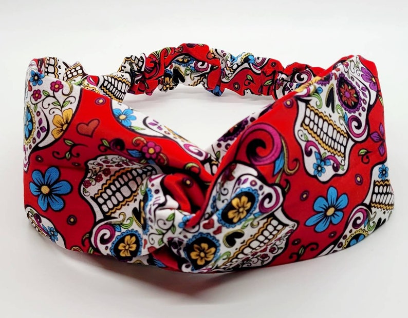 May include: Red headband with a white and colorful sugar skull pattern. The headband is made of a stretchy fabric and has a knotted design.