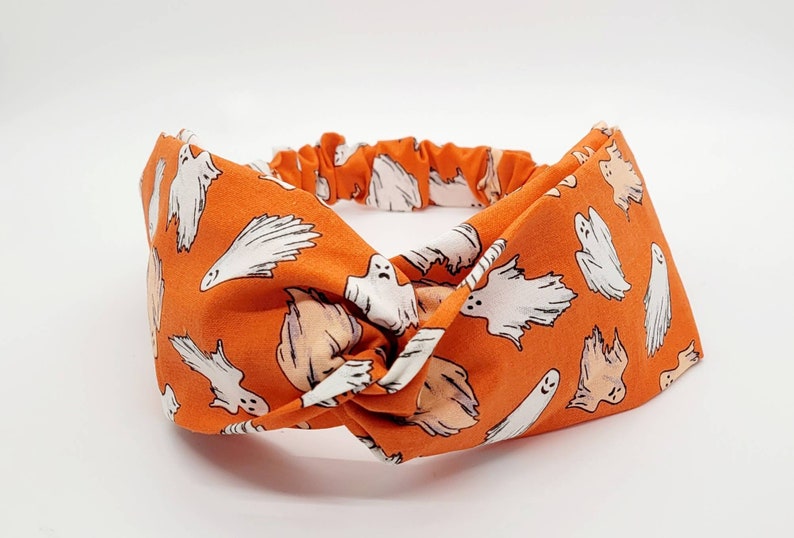 May include: Orange fabric headband with a white ghost pattern. The headband is knotted in the center.