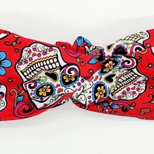 May include: Red headband with a white and black sugar skull pattern. The skulls are decorated with colorful flowers and swirls.