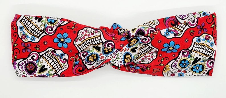 May include: Red headband with a white and black sugar skull pattern. The skulls are decorated with colorful flowers and swirls.