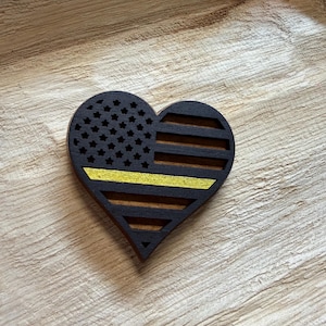 May include: Wooden heart-shaped decoration featuring a black and yellow American flag design. The flag includes stars and stripes, with a yellow stripe. The heart is made of dark wood and has a cutout design.