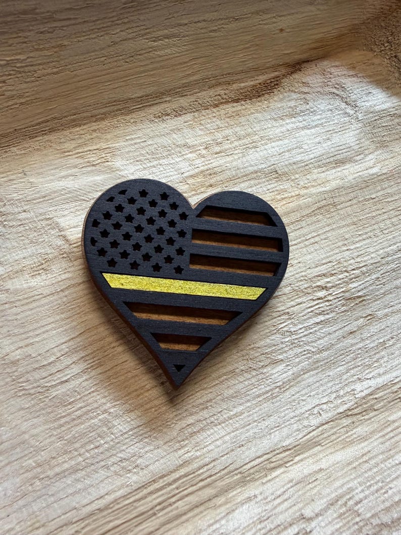 May include: Wooden heart-shaped decoration featuring a black and yellow American flag design. The flag includes stars and stripes, with a yellow stripe. The heart is made of dark wood and has a cutout design.