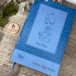 May include: A blue hardcover Holy Bible with a dotted pattern and a bookmark. The cover features an illustration of a child and the words "Holy Bible." The name "Easton Jayce Astacio" is printed on the bottom. A wooden Faith-Flame Engraving logo is visible.