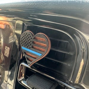 May include: A heart-shaped car air vent clip featuring a wooden design with the American flag and a blue line. The clip is made of wood and is attached to a car vent.