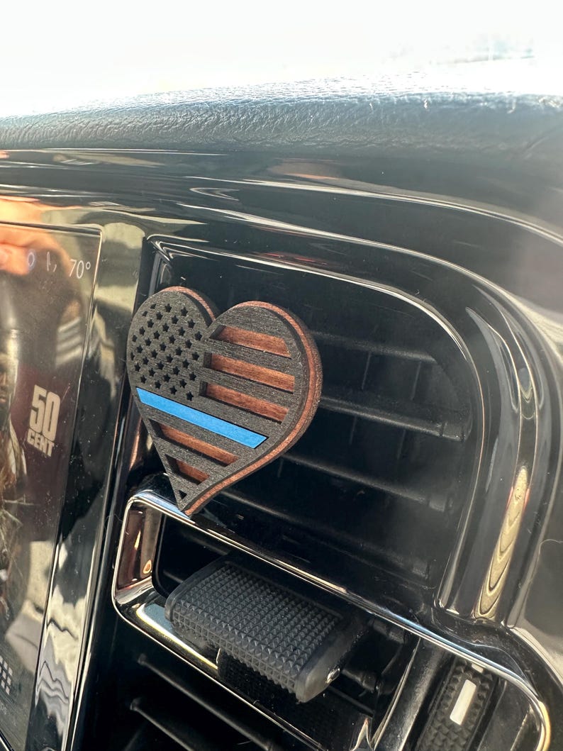 May include: A heart-shaped car air vent clip featuring a wooden design with the American flag and a blue line. The clip is made of wood and is attached to a car vent.