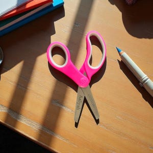 May include: Bright pink scissors with white handle inserts, resting on a wooden surface. The scissors have the name "Aaliyah" engraved on one blade and the brand "Westcott" on the other. A blue pencil and a stack of colorful papers are in the background.