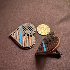 May include: Two heart-shaped wooden air fresheners with a patriotic design. The design features a blue line, a section of stars, and vertical stripes. A quarter is next to the air fresheners.