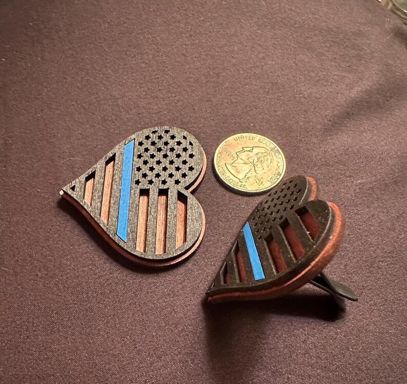 May include: Two heart-shaped wooden air fresheners with a patriotic design. The design features a blue line, a section of stars, and vertical stripes. A quarter is next to the air fresheners.