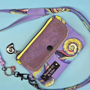 May include: A lavender lanyard wallet featuring a repeating yellow and blue spiral shell pattern. The wallet has a dark purple cork flap with a snap closure, a matching lavender tassel, a silver shell charm, and a black tag reading Wearable Art By Edye.