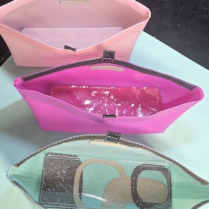 May include: Three open, rectangular pouches in shades of pink and clear. The top pouch is light pink, the middle is hot pink, and the bottom is clear. Each pouch has a contrasting interior and a rectangular item inside. The text "Wearable Art By Edge" is visible.