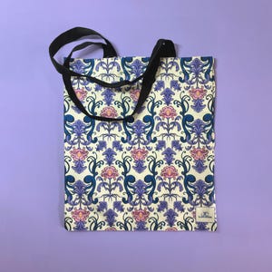 May include: Tote bag featuring a cream-coloured background with a repeating floral pattern in blue, purple, and pink. It has black handles and a small white tag with text. The bag is set against a purple backdrop.