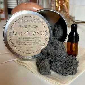 Op de afbeelding: Een ronde metalen doos met een zwart deksel dat donkergrijze vulkanische lavstenen bevat. De doos heeft een label met de tekst "Volcanic Lava Rocks Sleep Stones Powerless, Waterless, Flameless Room Diffuser" met gebruiksaanwijzingen. Naast de doos staat een klein bruin glazen flesje met een druppelaar.