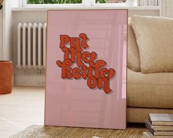 Peut inclure: Une affiche rose et orange avec le texte "Put the kettle on" dans une police de caractères rétro. L'affiche est accrochée à un mur blanc dans un salon.