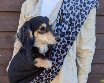 May include: A person wearing a grey and black leopard print dog carrier with a black dachshund inside. The carrier has a black strap and the text "One Paw Co." on it.
