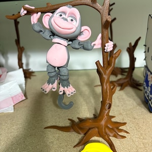 May include: A gray and pink monkey figurine hanging from a brown tree branch. The monkey has a smiling face and is holding onto the branch with its hands. A yellow banana rests at the base of the tree.
