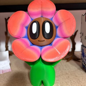 May include: A 3D-printed flower-shaped figurine with a brown face, large white eyes, and a smiling expression. The flower petals are orange and pink, with blue accents. The figurine has a green body and is standing upright.