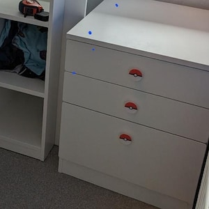 Puede incluir: Un conjunto de muebles blancos que incluye un armario, estanterías abiertas y una cómoda de tres cajones. Las puertas y cajones tienen pomos redondos con diseño de Poké Balls rojas y blancas. Una cinta métrica negra descansa sobre un estante.