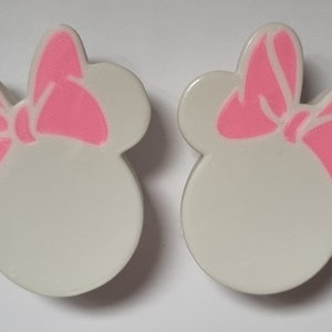 以下が含まれることがあります： ピンクのリボンが付いた、ミニーマウスの頭の形をした白いイヤリング2つ。デザインはシンプルで漫画的で、ファンに最適です。リボンは鮮やかなピンク色で、全体的な美しさはすっきりとしていてモダンです。