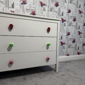 May include: A white three-drawer dresser featuring decorative drawer knobs. The top knobs are red Spider-Man masks, the middle knobs are green Hulk fists, and the bottom knobs are circular Captain America shields with a blue, white, and red design.