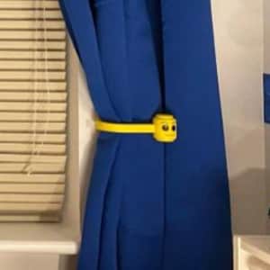 Könnte beinhalten: Blaue Vorhänge, die von einem gelben Lego-Vorhanghalter in Form eines Lego-Figurenkopfes gehalten werden. Links sind ein weißer Fensterrahmen und Jalousien zu sehen. Die Vorhänge hängen vor einer weißen Wand.