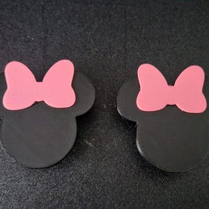 May include: Two black, three-dimensional mouse-shaped emblems, each featuring a smooth, matte finish and a prominent, solid pink bow attached to the top centre. The shapes have a distinct, layered texture visible on the surface of the black material.