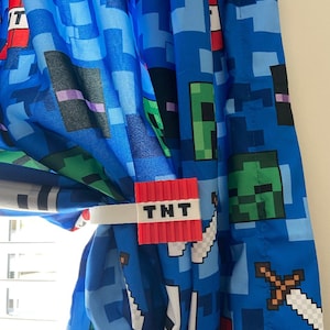 Könnte beinhalten: Ein roter und weißer Minecraft TNT-Vorhang-Halter. Der Halter ist in Form eines TNT-Blocks geformt, auf dem das Wort "TNT" aufgedruckt ist. Der Halter ist an einem blauen Vorhang mit einem Minecraft-Muster befestigt.