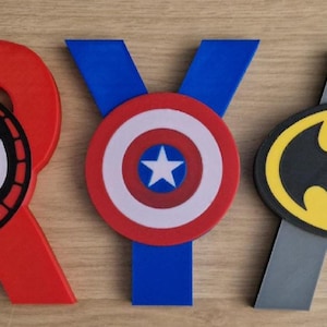May include: Wooden letters spelling "ARYAN" with superhero emblems. The letter "A" is purple with a Hulk fist, "R" is red with a Spider-Man face, "Y" is blue with Captain America's shield, "A" is gray with a Batman logo, and "N" is yellow with an Iron Man mask.