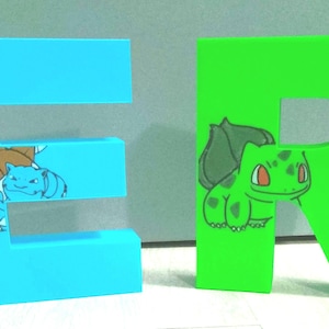 Puede incluir: Dos letras grandes e independientes, "E" y "R", en colores brillantes. La letra "E" es azul claro con una tortuga de dibujos animados y una criatura azul más pequeña. La letra "R" es verde con una criatura vegetal de dibujos animados.