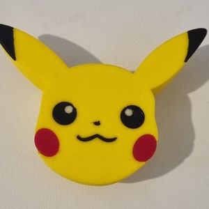 Puede incluir: Goma de borrar de goma amarilla con forma de Pikachu, un personaje de dibujos animados popular. La goma tiene ojos negros, una nariz negra y mejillas rojas.