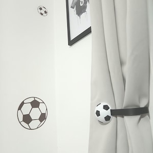 Pode incluir: Uma parede branca com adesivos de bola de futebol e uma impressão emoldurada de uma camisa com o nome "HARRISON" e o número "8". Uma cortina é presa por uma tira preta com um detalhe de bola de futebol.