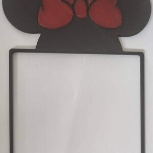 Op de afbeelding: Een zwart vierkant frame met een silhouet van muizenoren aan de bovenkant, geaccentueerd met een opvallende rode strik in het midden tussen de oren. Het frame heeft een dunne, platte rand en een hol midden, gemaakt van mat materiaal.
