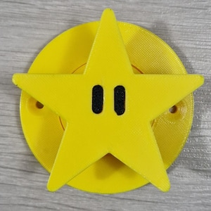May include: A bright yellow star with two black oval eyes, set on a yellow circular base. The star has five points and is a solid yellow colour. The base has a textured surface and small holes. This item is likely a decorative piece or a toy.