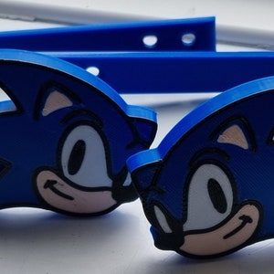 Könnte beinhalten: Zwei blaue Silikon-Armbänder mit jeweils einem Sonic the Hedgehog-Charakter-Anhänger. Die Anhänger sind blau und weiß mit schwarzen Umrissen.