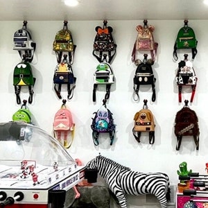 May include: A white wall with a collection of colorful mini backpacks hanging on hooks. The backpacks feature various cartoon characters, including Mickey Mouse, Minnie Mouse, and Stitch.