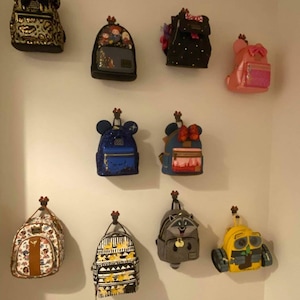 Könnte beinhalten: Eine Wanddekoration mit zehn Mini-Rucksäcken mit verschiedenen Disney-Charakterdesigns. Die Rucksäcke hängen an Haken und beinhalten Charaktere wie Mickey Mouse, Minnie Mouse, Stitch und andere.