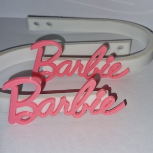 May include: Two pink plastic Barbie nameplates, each with the word "Barbie" written in a cursive font. The nameplates are on top of a white plastic headband.