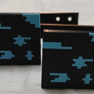 Könnte beinhalten: Zwei schwarze Leder-Gürtelschlaufen mit quadratischen, schwarzen Mittelstücken. Jedes Mittelstück hat ein blaues, pixeliges Design. Die Gürtelschlaufen sind verbunden.