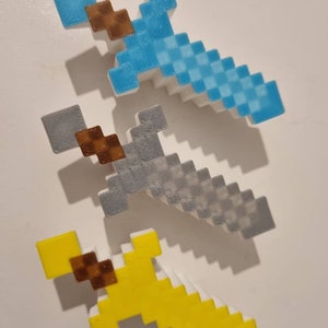 Puede incluir: Tres espadas de Minecraft hechas de plástico. Las espadas son azules, grises y amarillas. Cada espada tiene un mango marrón.
