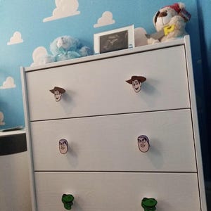 May include: White chest of drawers with four drawers featuring Toy Story character themed drawer pulls. The top drawer has Woody, the second drawer has Buzz Lightyear, the third drawer has Mr. Potato Head, and the bottom drawer has Rex the dinosaur.